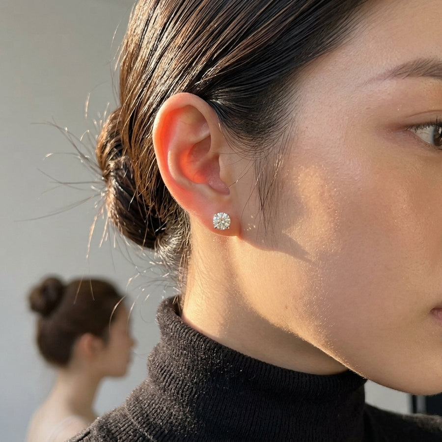 a close-up of a woman's ear wearing a small round 1 ct stud earring with a clear gemstone, set against a softly blurred background.