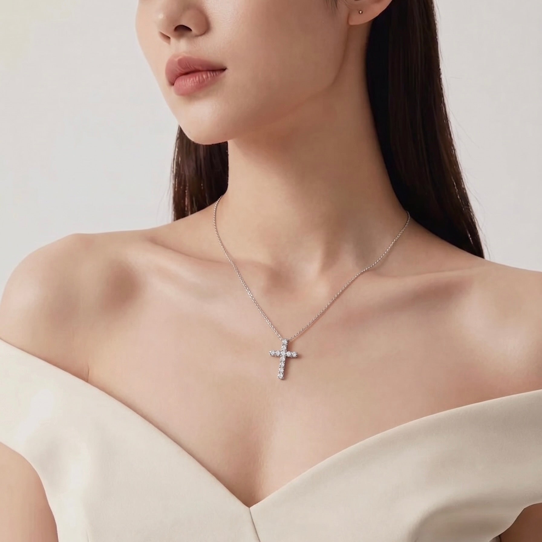 A close-up of a woman wearing a delicate 14k gold  cross pendant necklace adorned with 0.2ct round diamonds, paired with a soft off-the-shoulder cream top against a light neutral white background.